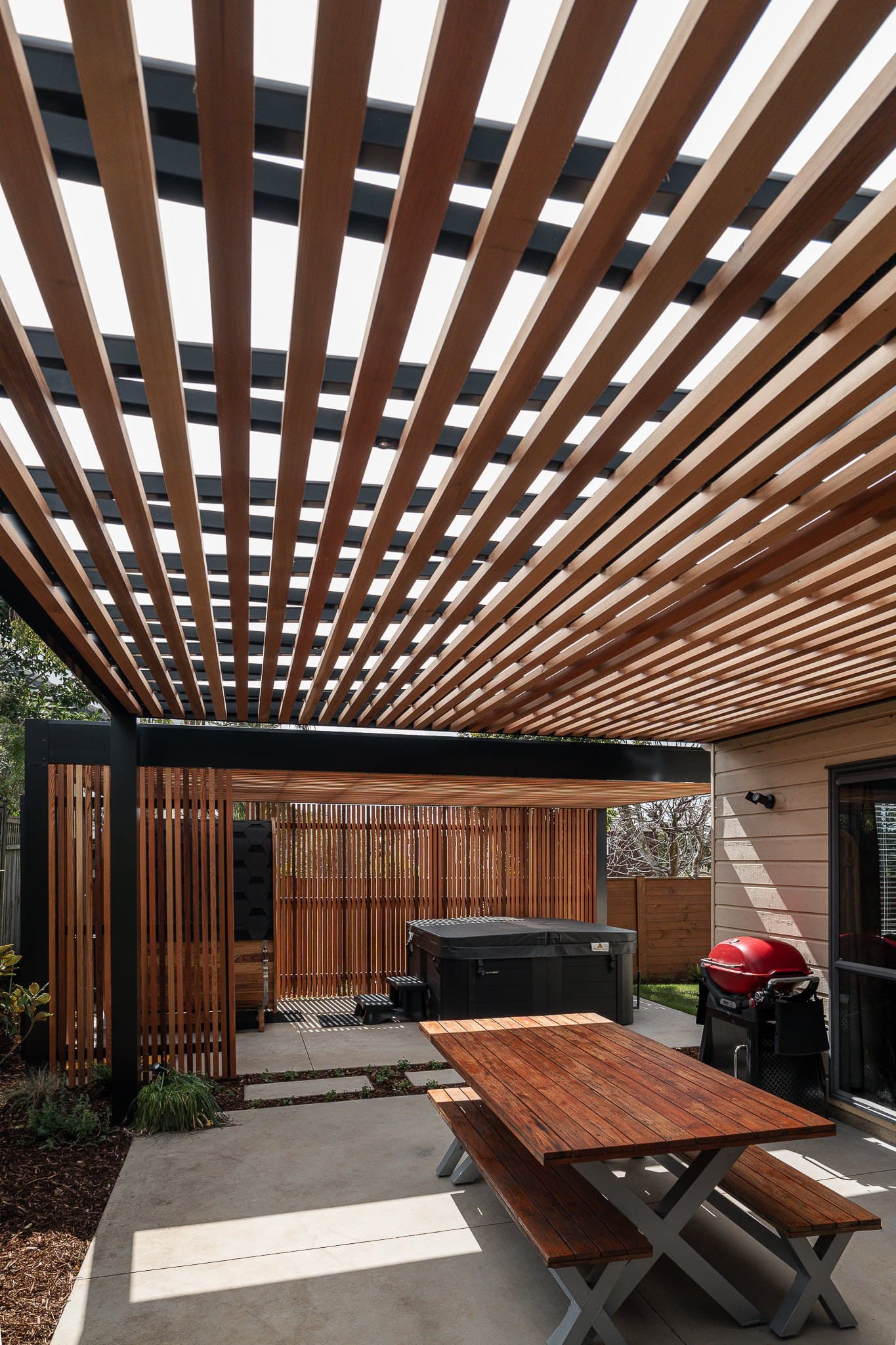 Outdoor dining table beneath cedar roof battens, looking towards the screened spa area